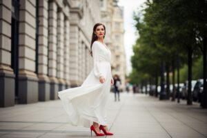 žena v bielych šatách a červených lodičkách stojí na mestskom chodníku s rozmazaným pozadím
